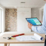 Un homme examine des plans numériques et des coûts sur une tablette, devant des plans architecturaux et outils de mesure dans une pièce en rénovation.