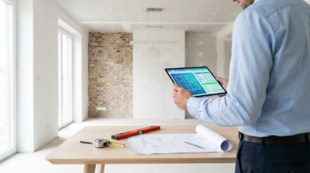 Quel est le prix travaux m2 en 2026 selon votre projet ? Un homme examine des plans numériques et des coûts sur une tablette, devant des plans architecturaux et outils de mesure dans une pièce en rénovation.